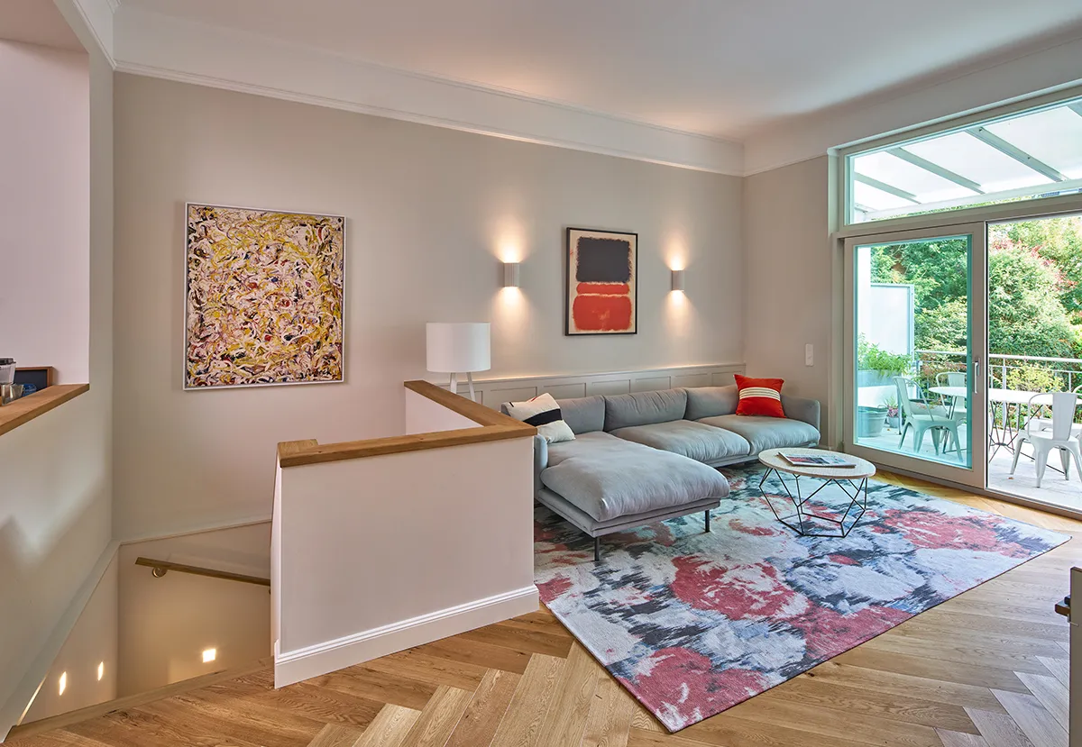 Wohnbereich einer Maisonettewohnung mit großer Fensterfront, moderner Ecksofa-Lösung, farbigem Teppich und Zugang zur überdachten Terrasse.